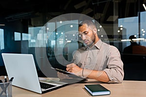 Serious concentrated businessman writing document while sitting in office using laptop for online learning, hispanic man