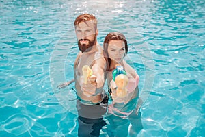 Serious and concentrated couple stands back to back in swiming pool and look on camera. They pose. Guy and girl holds