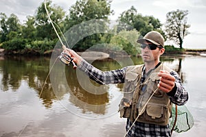 Serious and concentrated man stands in water and holds fly-fishing with reel under it in one hand and part of spoon in