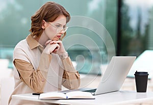 Serious concentrated young female office worker working remotely on laptop online sitting outdoors