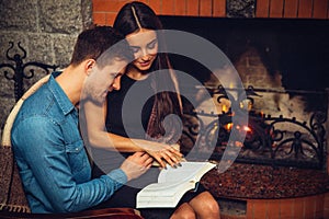 Serious and concentrated young man and woman sit together at fireplace. They read opened book. Model touch page with