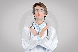 Serious confident man doctor in white coat crossed his arms and making stop gesture, posing on gray background