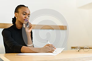 Serious confident young African or black American business woman on phone taking notes in office