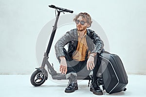 Serious and curly man wearing sunglasses posing in the bright studio while sitting on the scooter with a backpack