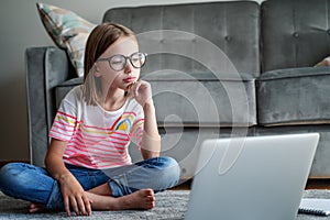 Serious cute little girl 8 years old in a striped T-shirt and jeans with glasses sits at home on a carpet in front of a laptop,