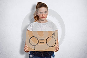 Serious cute young feminist with pony tail, holds plate with sign which demonstrates quality between sexes, dressed in casual t sh