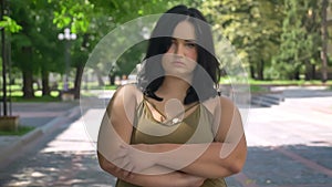 Serious depressed young pretty woman with obesity looking at camera, standing on street in park during sunny day