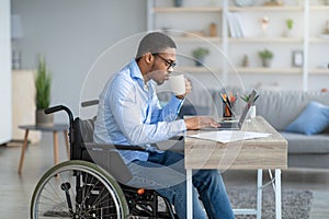 Serious disabled black man in wheelchair drinking coffee, watching video lesson on laptop, working online from home