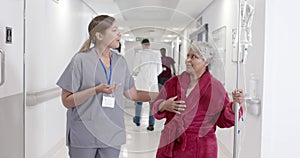 Serious diverse female nurse talking to senior patient with drip walking in slow motion, unaltered
