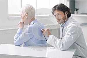 Serious doctor listening to coughing senior patient at clinic