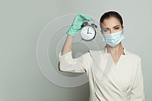 Serious doctor woman in medical protective face mask and professional uniform holding alarm clock on white background