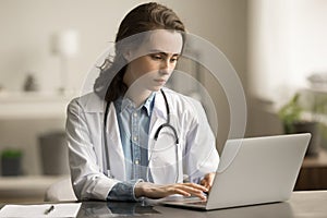 Serious doctor woman in white uniform coat typing on laptop