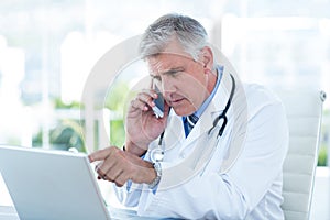 Serious doctor working on laptop and having phone call