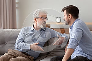 Serious elderly father and grownup son sitting on couch talking