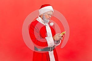 Serious elderly man with gray beard in santa claus costume using smart phone with concentrated