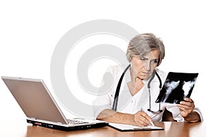 Serious elderly woman doctor sitting at table with computer and looking at x-ray