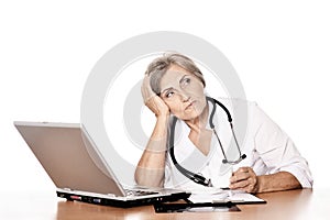 Serious elderly woman doctor sitting at table with computer on white