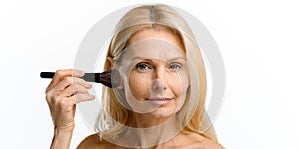 Serious elegant beautiful middle-aged blonde woman applying make-up with brush isolated on white, headshot of mature