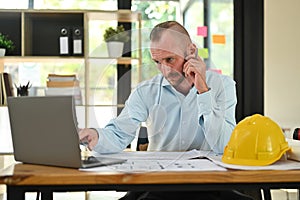Serious engineer man watching online webinar training, checking construction plan on laptop at his workstation