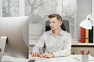 Serious and engrossed business man in shirt sitting at the desk, working at computer with modern monitor. Manager or worker.