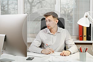 Serious and engrossed business man in shirt sitting at the desk, working at computer with modern monitor. Manager or worker.