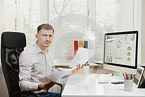 Serious and engrossed business man in shirt sitting at the desk, working at computer with modern monitor. Manager or worker.