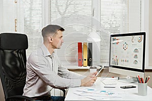 Serious and engrossed business man in shirt sitting at the desk, working at computer with modern monitor. Manager or worker.