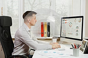 Serious and engrossed business man in shirt sitting at the desk, working at computer with modern monitor. Manager or worker.