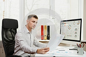 Serious and engrossed business man in shirt sitting at the desk, working at computer with modern monitor. Manager or worker.