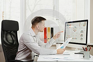 Serious and engrossed business man in shirt sitting at the desk, working at computer with modern monitor. Manager or worker.