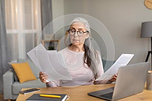 Serious european old female with gray hair in glasses works on computer and documents in room interior