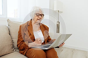 a serious, experienced, elderly woman is sitting working from home on the couch with a laptop on her lap and menacingly