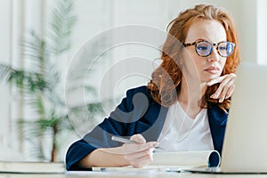 Serious experienced female coach makes records in notepad, focused at screen of laptop computer, has long red hair, wears