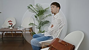 Serious-faced young hispanic man, an elegant professional, sitting pensively in a waiting room chair, casting concentrated glances