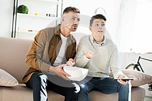 Serious father and teen son watching tv and holding bowl of popcorn