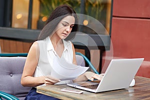 Serious female company owner concentrated into documents, reads attentively terms of contract, uses laptop computer, spends dinner