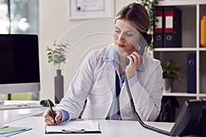 Serious Female Doctor Or GP Wearing White Coat Sitting At Desk In Office Making Phone Call