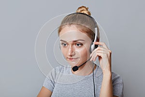 Serious female helpline operator uses headphones for her work, being focused on something, isolated over grey background. Business