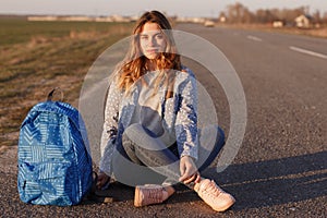 Serious female hitchhiker sits crossed legs on asphalt road with her bag, enjoys travelling in countryside, explores unknown place