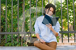 Serious female model focused on reading, sits at wall in the park. Concetrated student prepares for coming session at college.