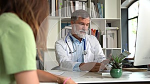 Serious focused aged physician in medical coat sitting at table, consulting female patient about illness or surgery