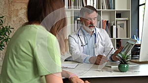 Serious focused aged physician in medical coat sitting at table, consulting female patient about illness or surgery