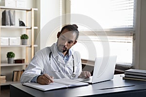Serious focused GP doctor writing medical records at open laptop