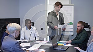 Serious focused male CEO passing paperwork to employees sitting at conference table in office. Portrait of confident