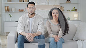 Serious focused unemotional multiethnic married couple family sitting on sofa in home living room interior, arabic