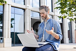 A serious and focused young man is sitting outside an office center on a bench, wearing headphones and holding a laptop