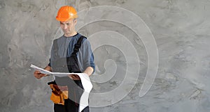a serious foreman examines the plan and blueprints standing against a concrete wall in an orange helmet and glasses