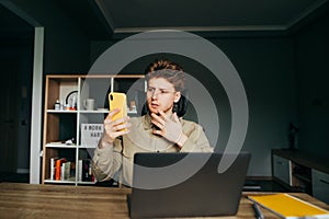 Serious freelancer sitting at home at work with a laptop, using a smartphone in a focused face. Concentrated young man in a shirt