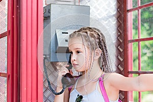 A serious girl with afro-braids makes a phone call in an old English telephone booth. Generational contrast