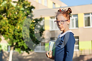 Serious girl in a glasses stands with classbook near a school